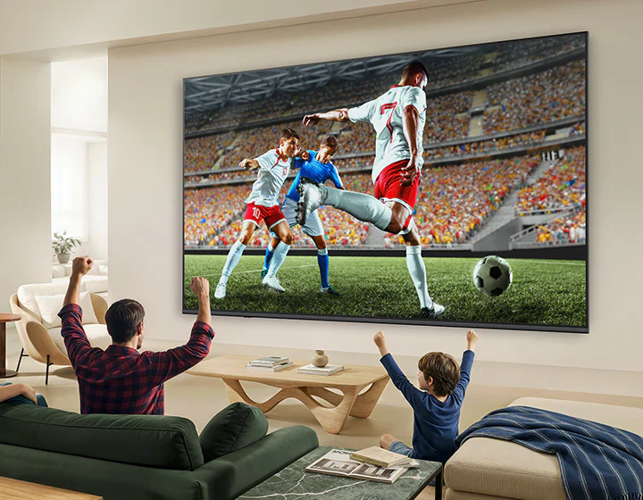 Телевизор LG QNED evo AI QNED82 Mini LED Ultra Big показывает захватывающую крупномасштабную футбольную игру с улучшенным качеством изображения и яркими цветами, а семья вместе радуется на диване в гостиной.